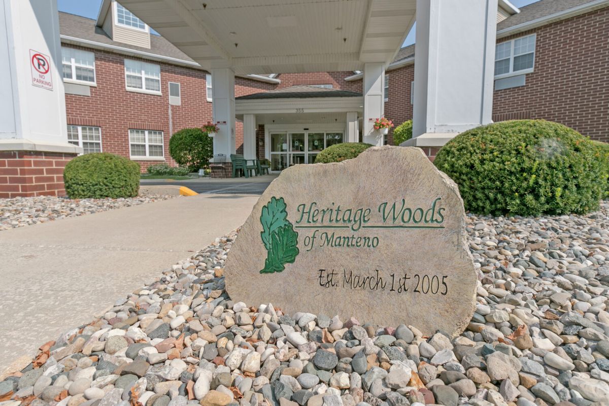 The entrance to a building with a sign on a stone, surrounded by landscaping.