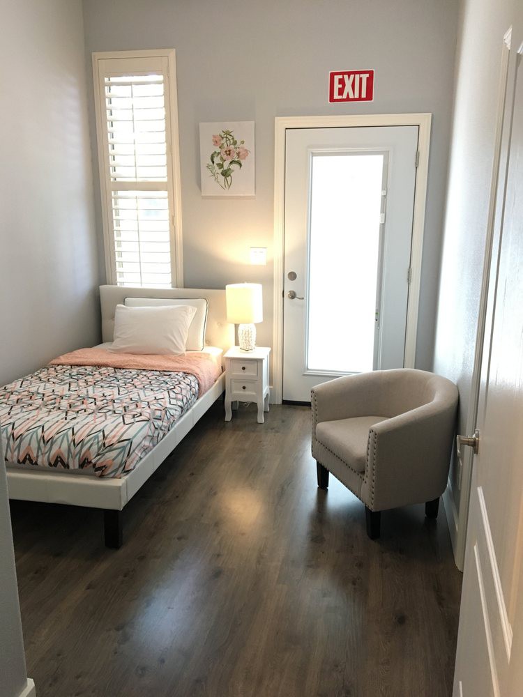 A cozy, minimalist bedroom featuring a single bed, armchair, and nightstand with a lamp.