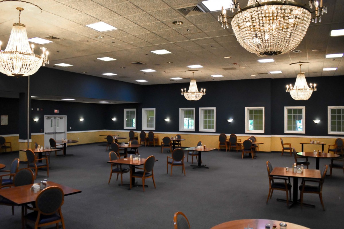An elegant banquet hall with chandeliers, neatly arranged tables and chairs, and multiple windows.