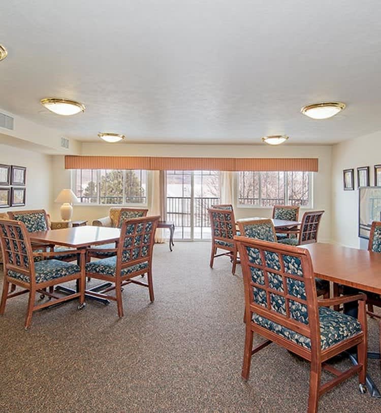 A well-lit communal living room with several tables and chairs, featuring large windows for natural light.