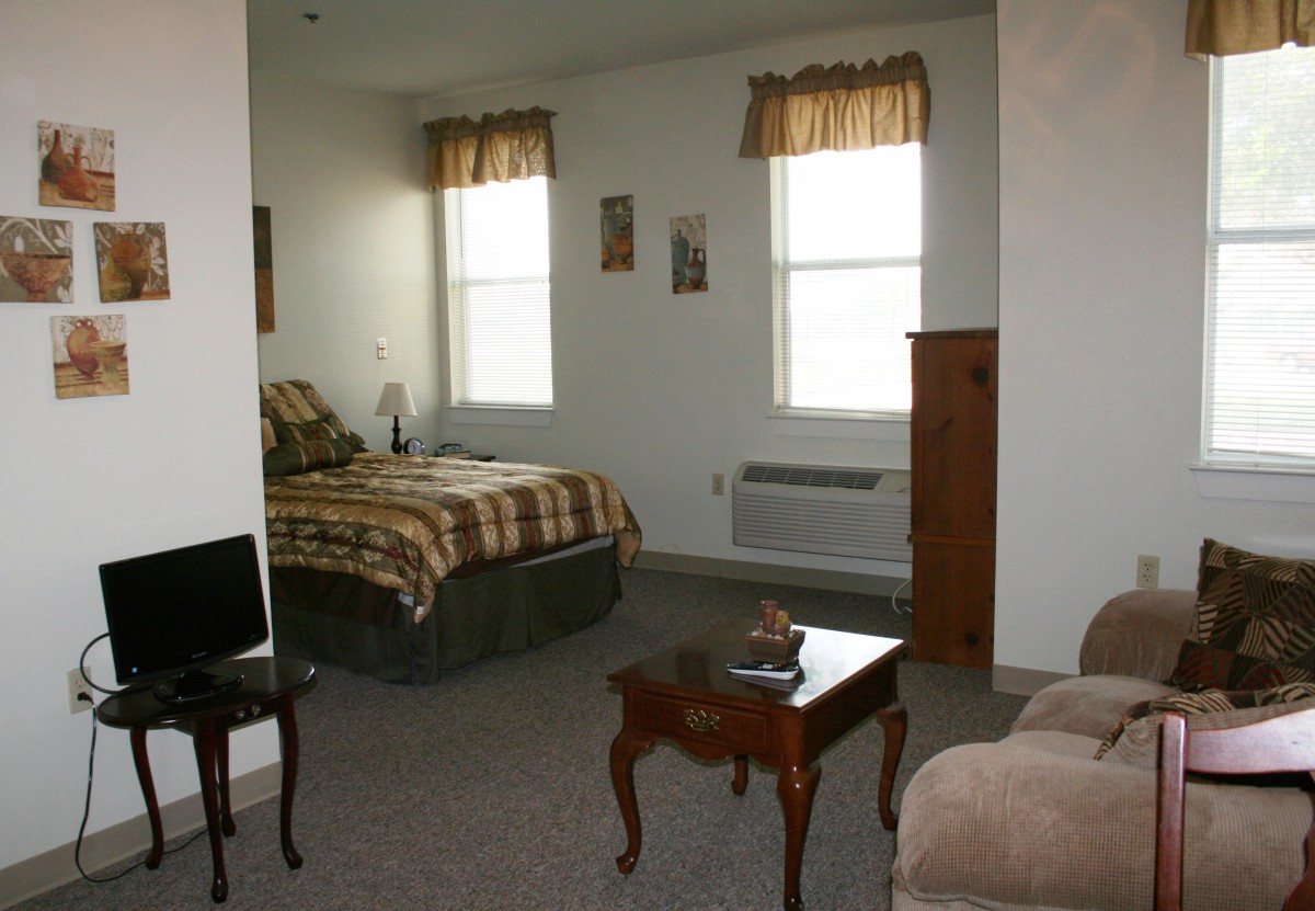 A cozy bedroom with a bed, small table, and a chair near two windows.