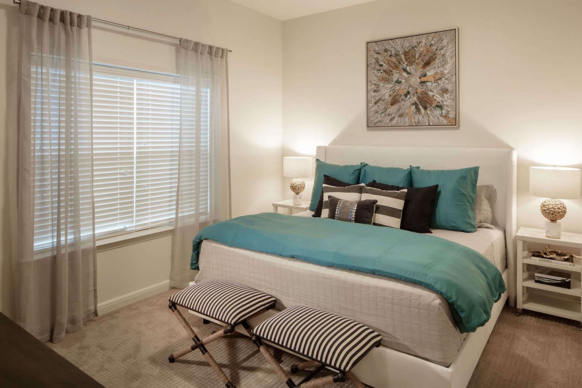 A modern bedroom with teal bedding, artistic wall decor, and ample natural light from a window with blinds.