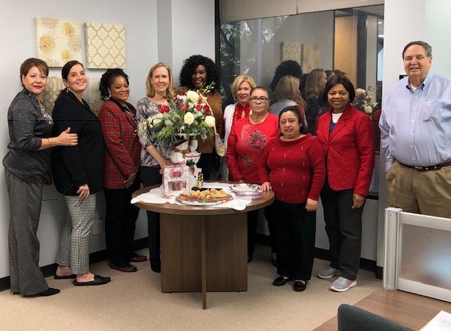A group of people in an office setting gathered around a table with flowers and desserts, likely celebrating an event.