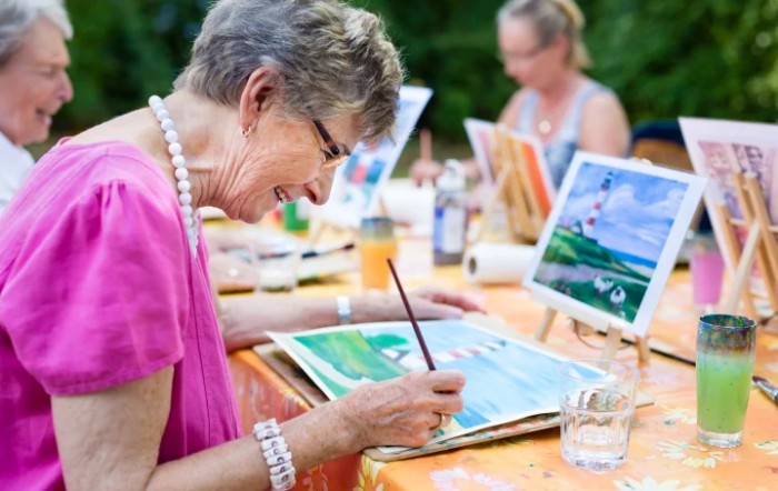 An elderly woman smiling as she paints during an outdoor art class.