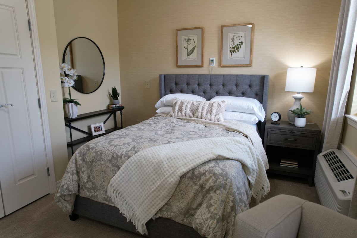 A cozy bedroom with a neatly made bed, decorative pillows, artwork on the walls, and a side table with a lamp.