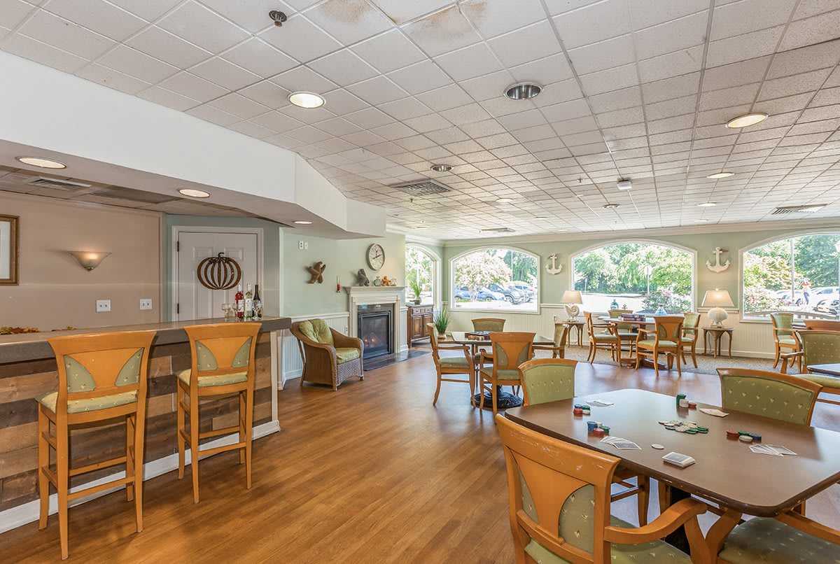 A spacious dining room with wooden flooring, featuring several tables and chairs, alongside a bar area filled with natural light.