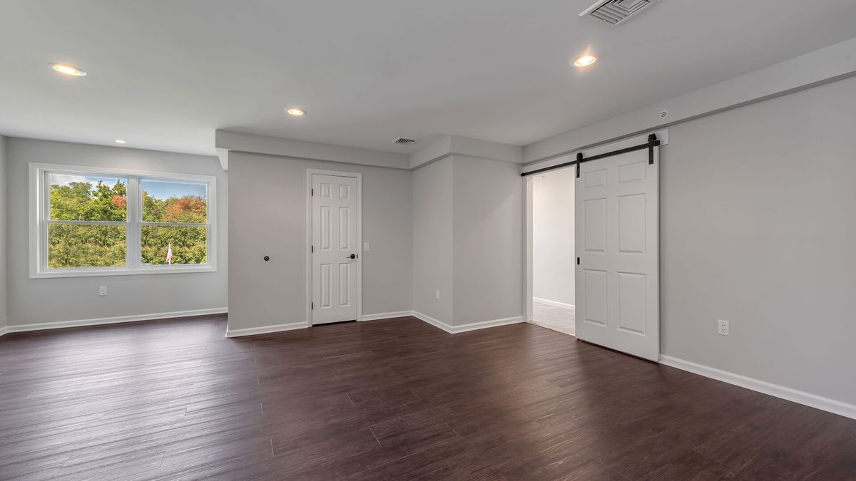 A modern, empty room with hardwood floors and white walls featuring a barn door and recessed lighting.
