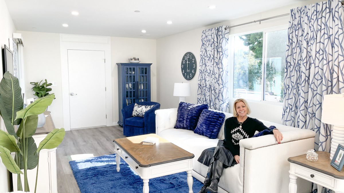 A well-lit living room featuring a white sofa with blue pillows, a matching blue rug, and an elegant armoire. A person is seated on the sofa.