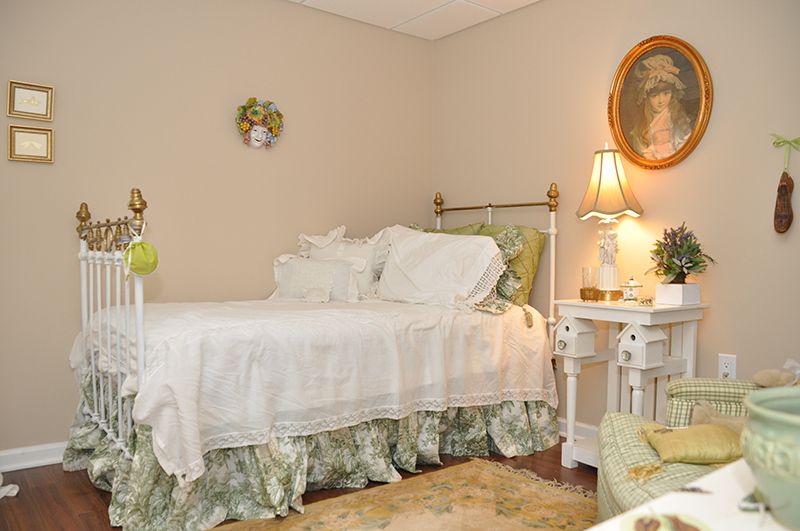 A cozy vintage-style bedroom with a single bed, adorned with white bedding and floral accents. A lamp and framed wall art add charm to the room.