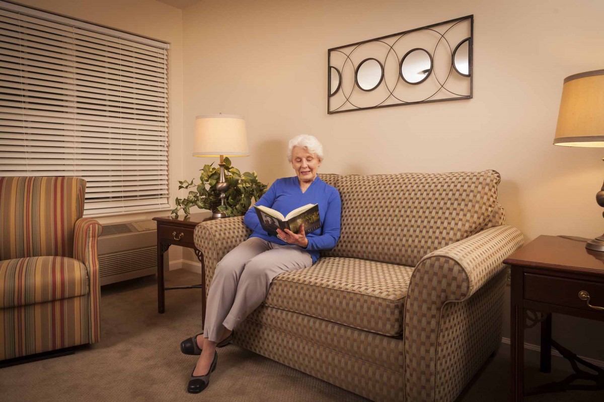 An elderly woman sitting on a sofa in the living room, reading a book with a warm lamp lighting the space.