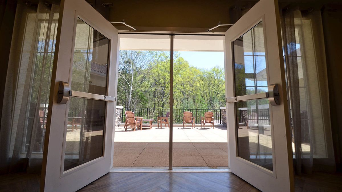 A view through open double doors revealing a sunny patio with outdoor seating and trees in the background.
