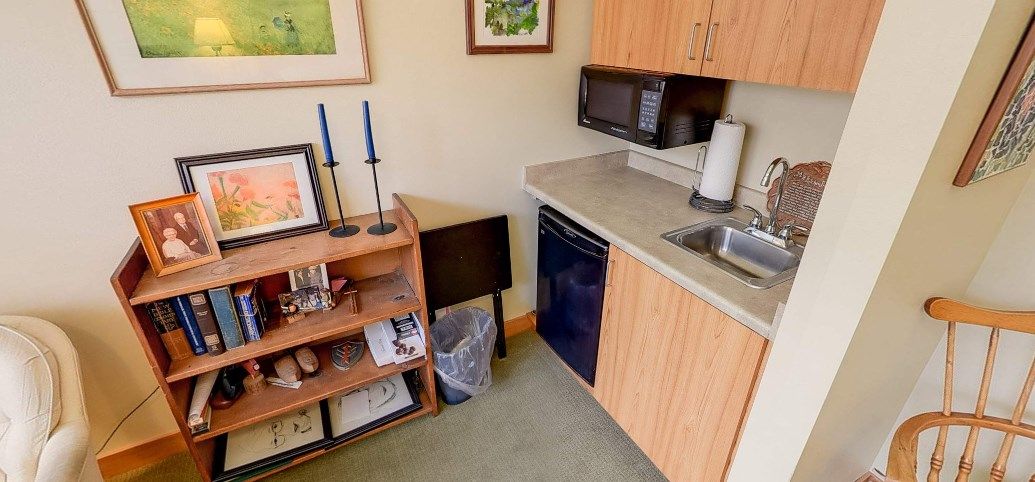 A compact kitchenette with a microwave, mini fridge, and wooden cabinets, alongside a bookshelf topped with candlesticks.