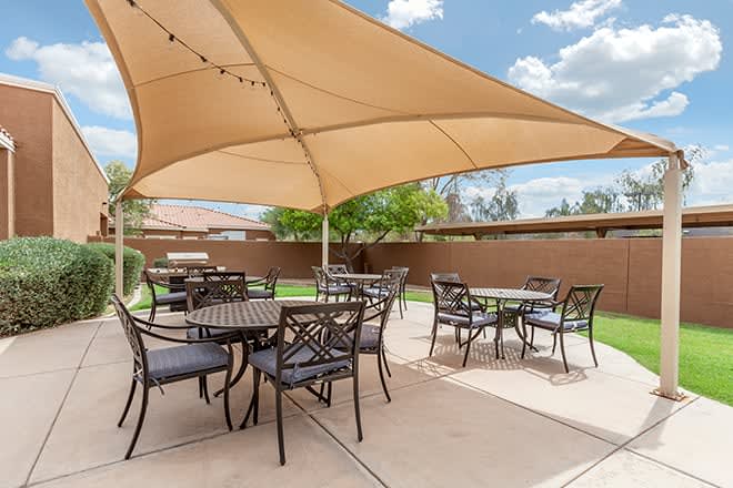A sunny outdoor patio with tables and chairs under a large shade sail.