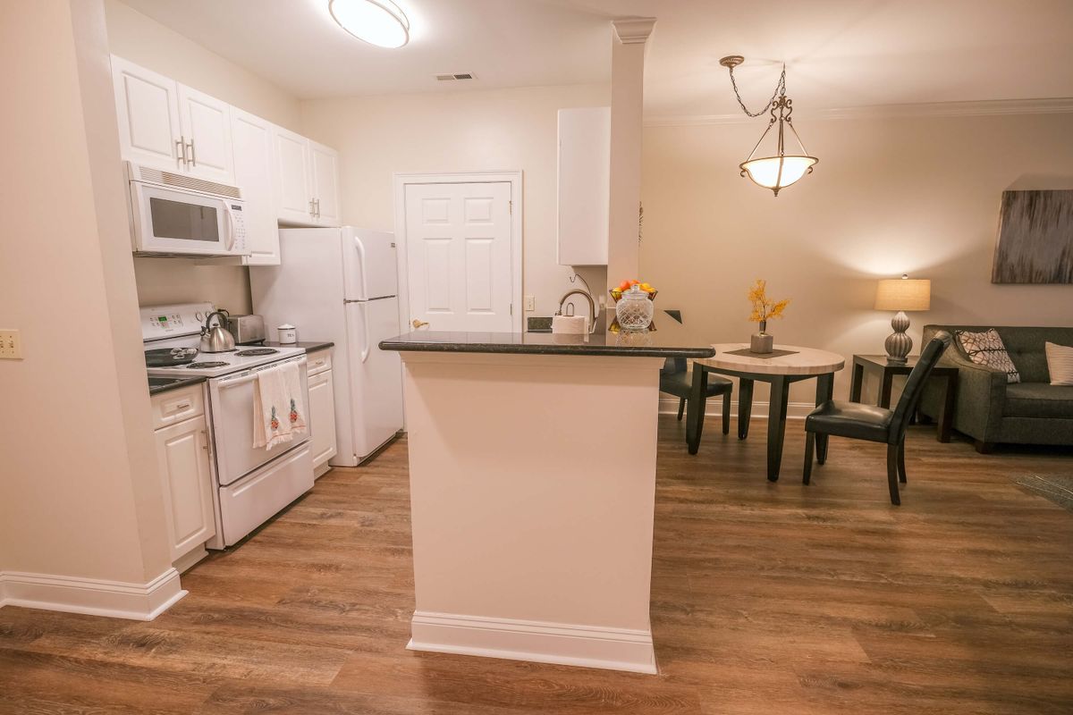 A modern apartment kitchen with white cabinets and a small dining area, featuring wood flooring and a chandelier.