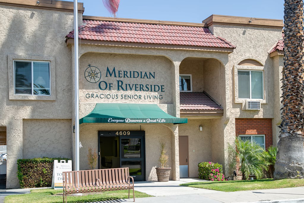 Entrance of the Meridian of Riverside senior living facility with an awning and landscaped surroundings.