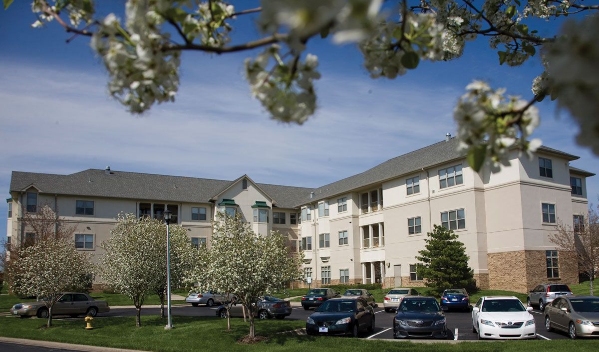 A residential apartment complex surrounded by blooming trees and a parking lot with cars under a cloudy sky.