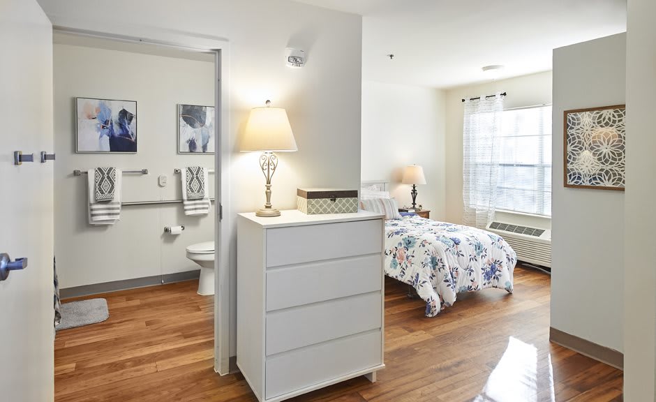 A cozy bedroom with a bed, dresser, and bedside lamp, featuring hardwood floors and a window letting in natural light.