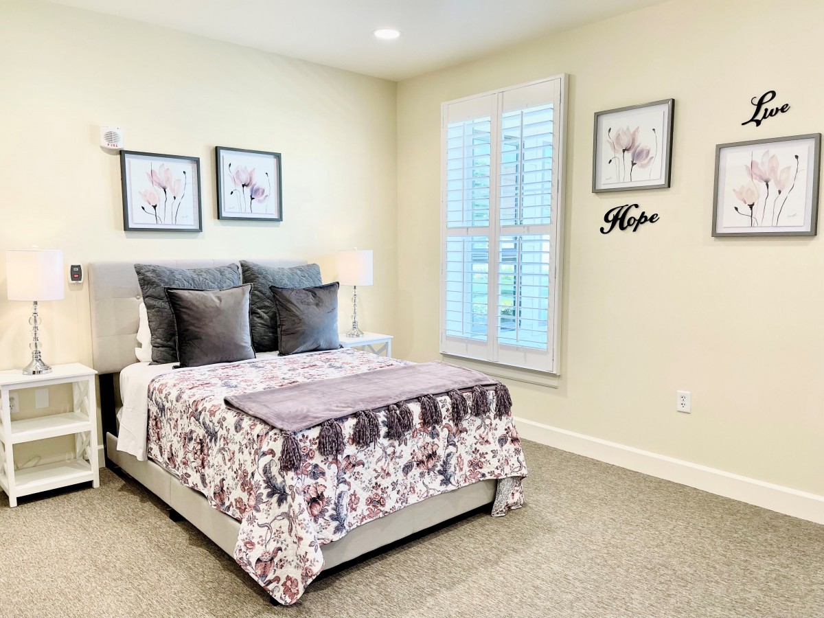 A cozy bedroom with a neatly made bed, adorned with decorative pillows and a floral blanket. The walls feature framed artwork, and a window allows natural light to fill the space.