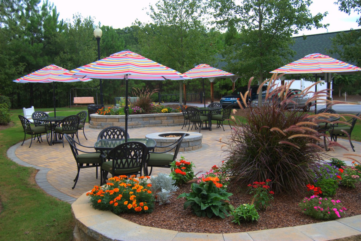 A well-landscaped outdoor patio area with tables, chairs, and umbrellas, surrounded by vibrant flowers and featuring a central fire pit.