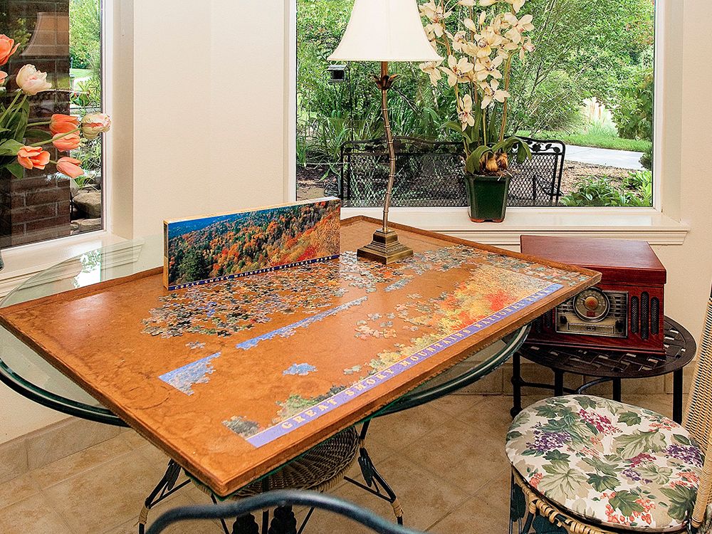 A jigsaw puzzle in progress on a wooden table near a lamp and window, surrounded by indoor plants.