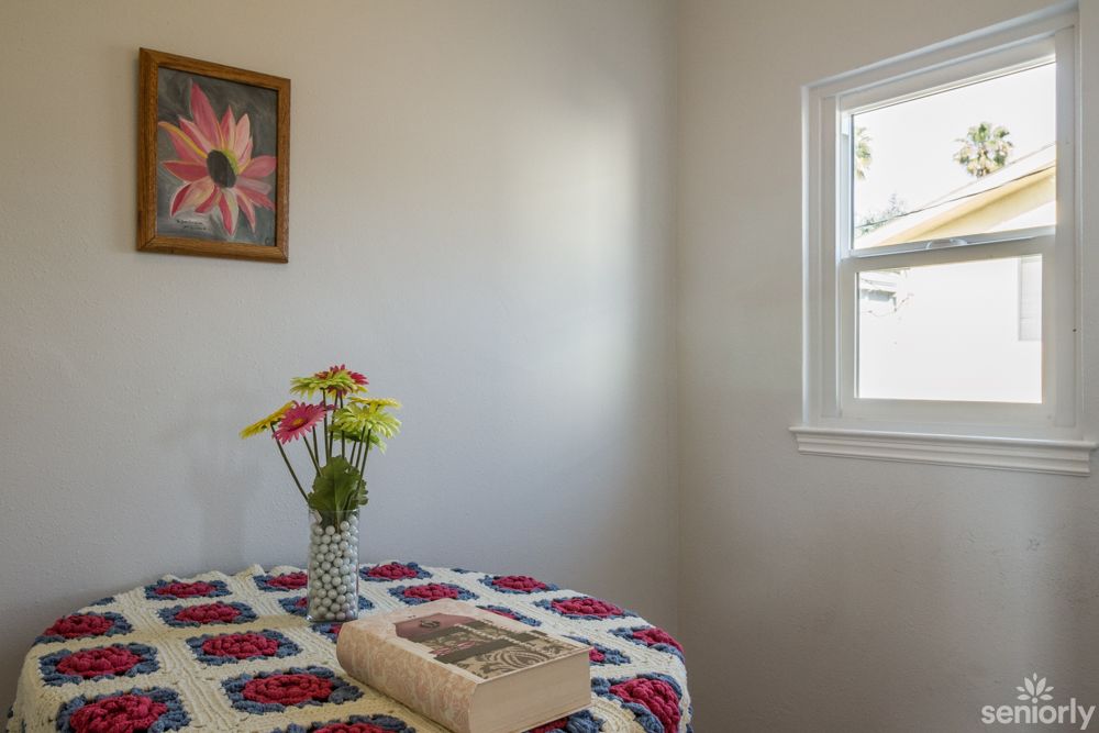 A cozy corner featuring a table with a floral quilt, topped with a vase of colorful flowers and an open book, next to a window.