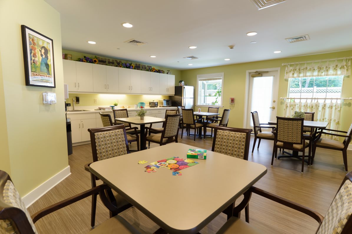 A well-lit dining and activity room with multiple tables and chairs, featuring a kitchenette area.