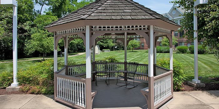 A wooden gazebo in a lush garden setting, featuring chairs inside and a well-manicured lawn in the background.