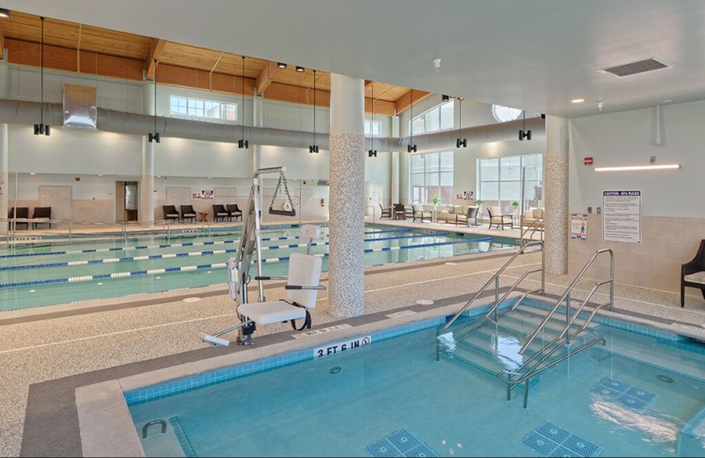 An indoor swimming pool area featuring a separate hot tub with accessibility equipment and seating around the pool.
