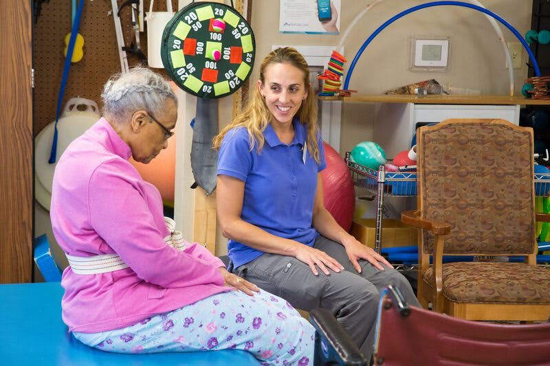 A female therapist assists an elderly woman with physical therapy exercises in a rehabilitation center.