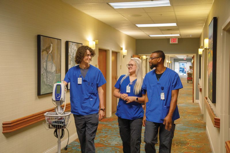 Three medical professionals in blue scrubs walk down a hospital corridor, engaging in conversation and showing teamwork.