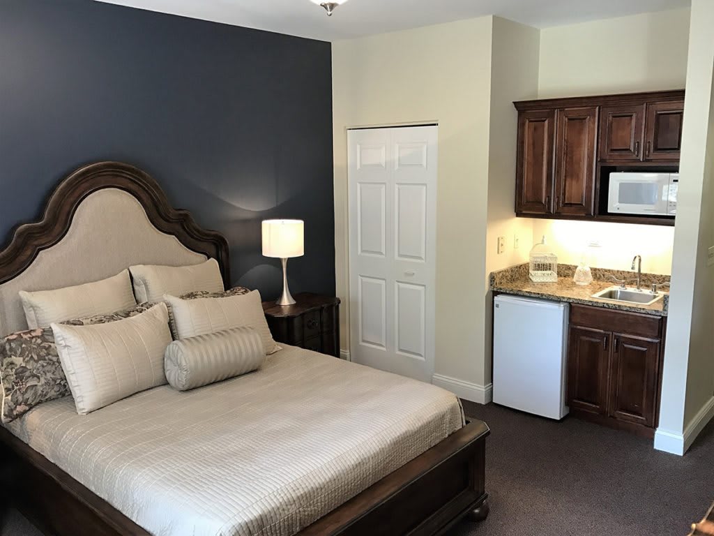 A cozy bedroom with a bed, lamp, and kitchenette.