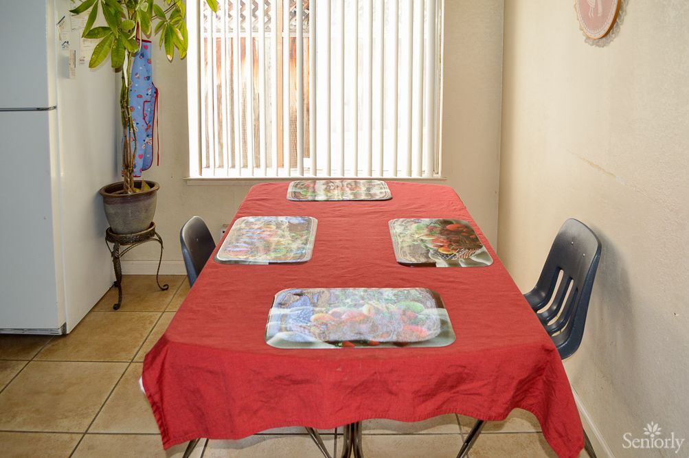 A dining table with a red tablecloth and placemats set for four, situated near window blinds and a potted plant.