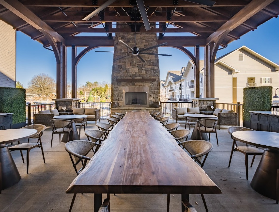 A modern outdoor dining area with a long wooden table, surrounded by chairs, and featuring a central fireplace.