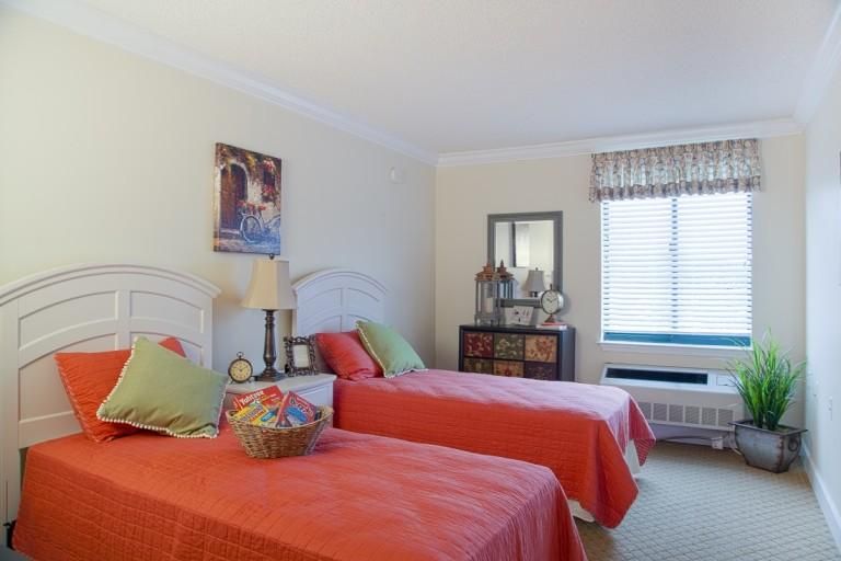 A cozy bedroom with two twin beds adorned with orange bedspreads, a window with blinds, and decorative elements enhancing the room's ambiance.
