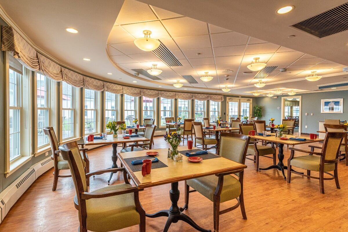 A cozy dining room with neatly arranged tables and chairs, featuring large windows and elegant lighting.