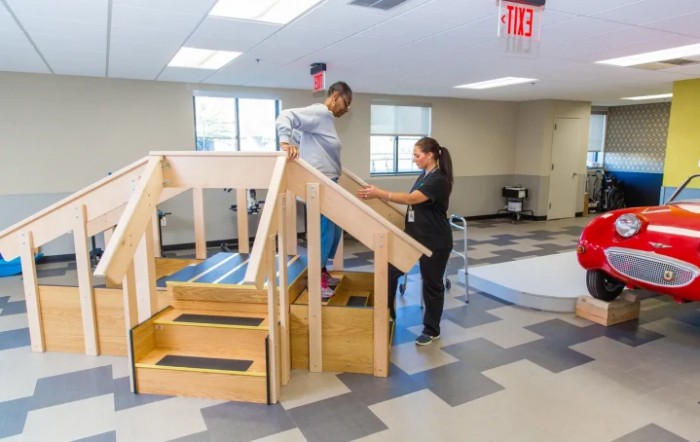A person receives assistance during physical therapy, practicing walking on stairs indoors.