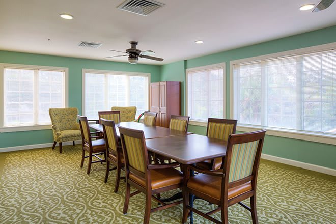 A well-lit dining room with a large wooden table and chairs, featuring green walls and ample natural light from surrounding windows.