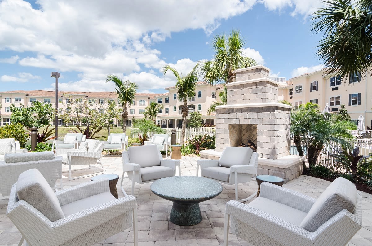 A stylish outdoor patio with modern furniture and a stone fireplace, surrounded by palm trees and buildings under a cloudy sky.