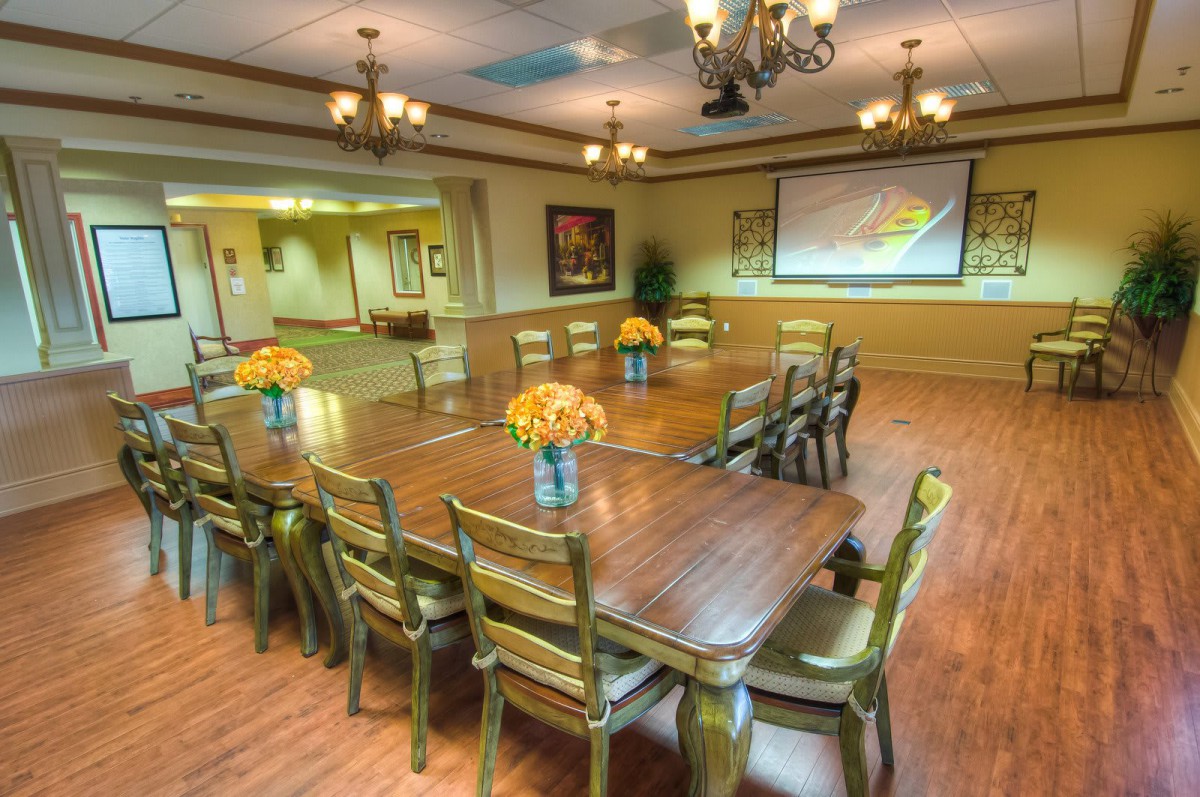 A well-lit conference room with a large table, surrounded by chairs, featuring a projector screen and floral centerpieces.