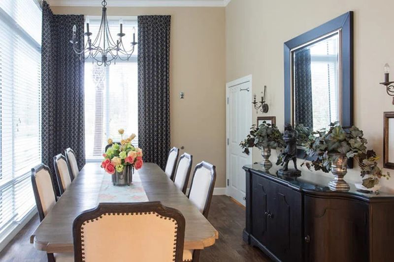 A stylish dining room featuring a long table with chairs, a chandelier, a vase of flowers, and large curtains.