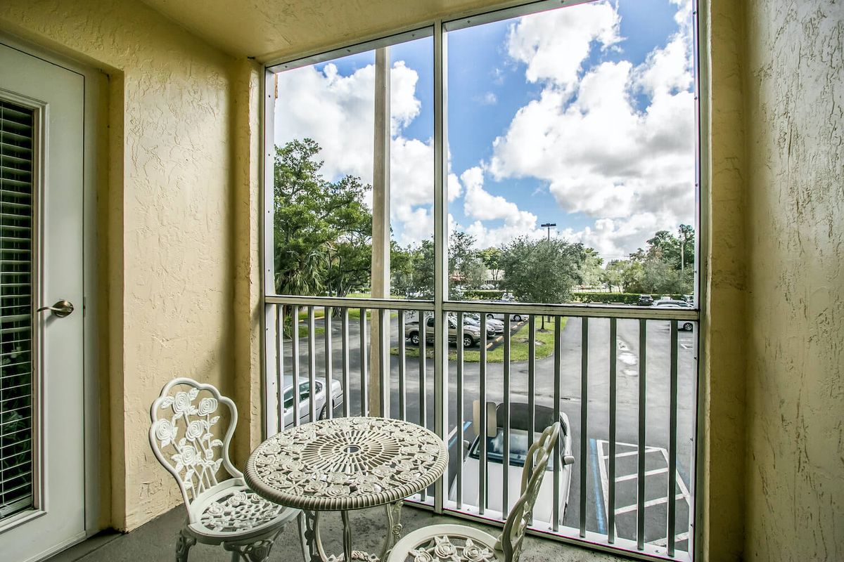 A quaint balcony with decorative chairs and a table, overlooking a parking area under a partly cloudy sky.
