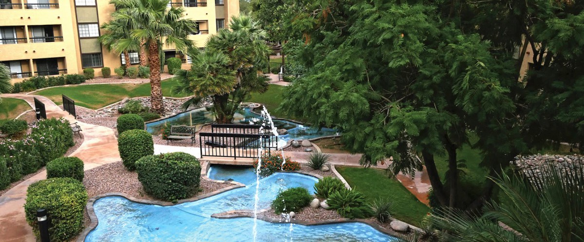 A scenic garden landscape featuring a small bridge over a water stream, surrounded by lush greenery and trees.