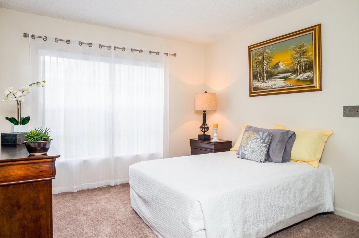 A cozy bedroom with a neatly made bed, decorative pillows, a side table with a lamp, and a landscape painting on the wall.