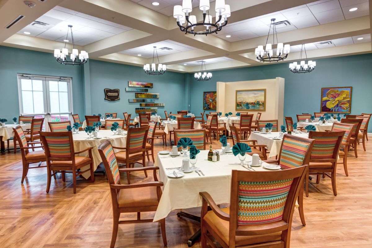 A well-decorated dining room featuring elegantly set tables, wooden chairs with colorful cushions, and chandeliers overhead.