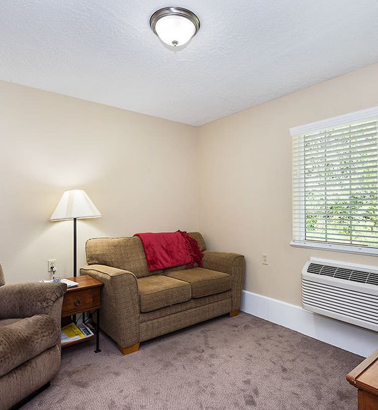 A cozy living room with a sofa, chair, and lamp, featuring a window and air conditioner.