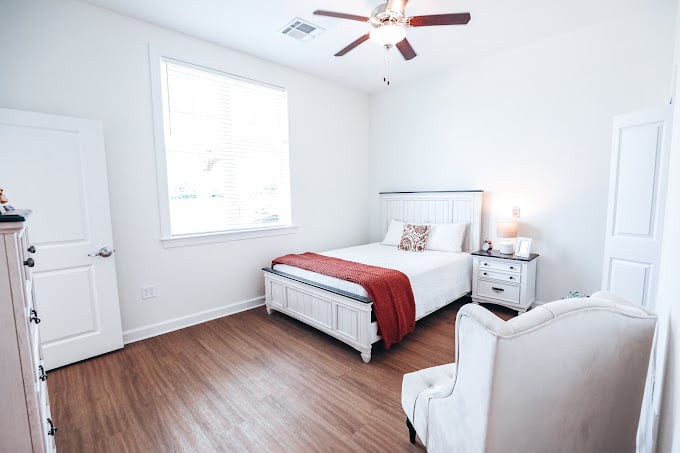 A bright and minimalist bedroom with wooden flooring, featuring a neatly made bed adorned with a red blanket and white furniture.