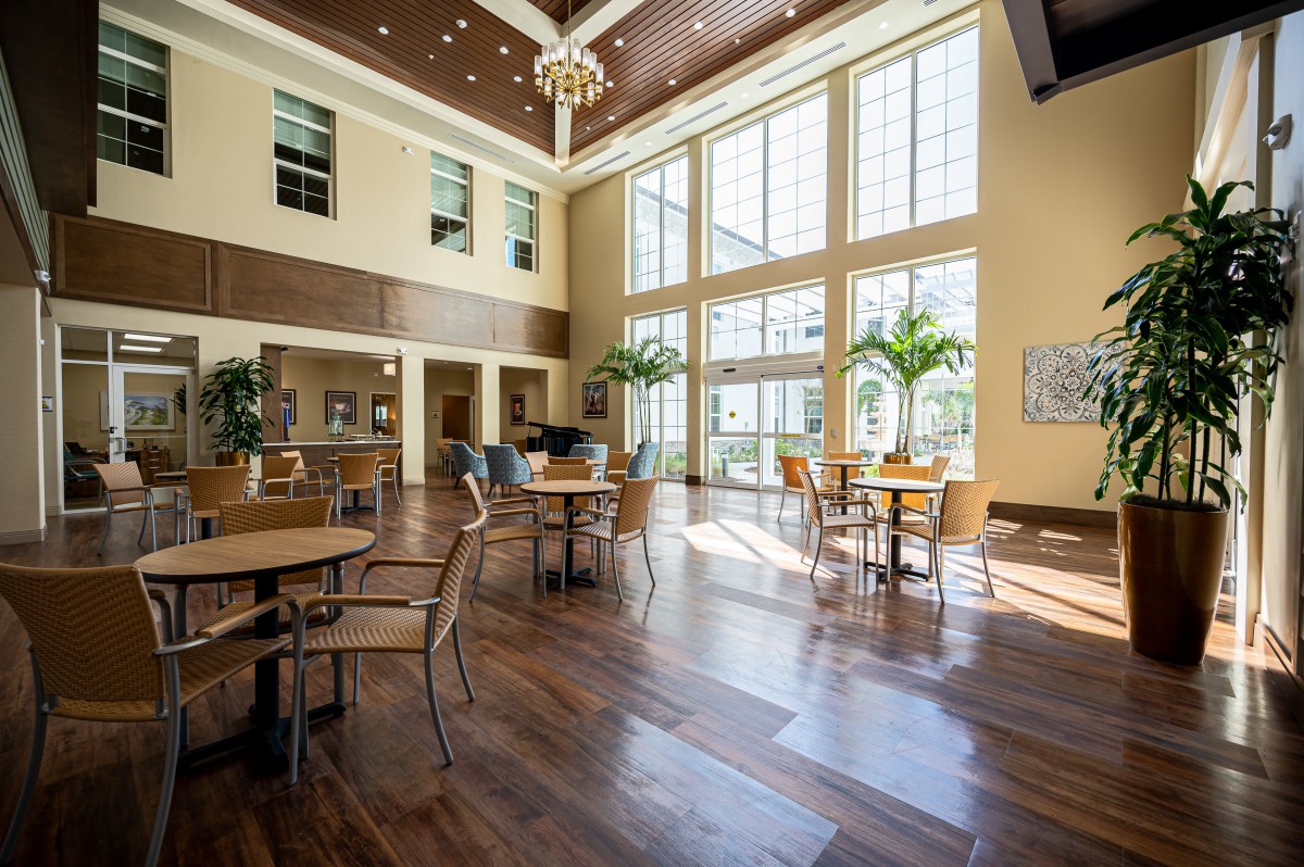 A spacious, sunlit lobby featuring wooden floors, chairs, and tables.