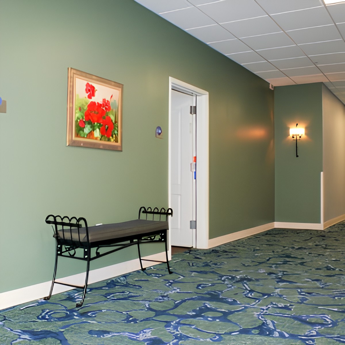 A hallway with a bench, painting, and door, featuring patterned carpet and wall sconces.