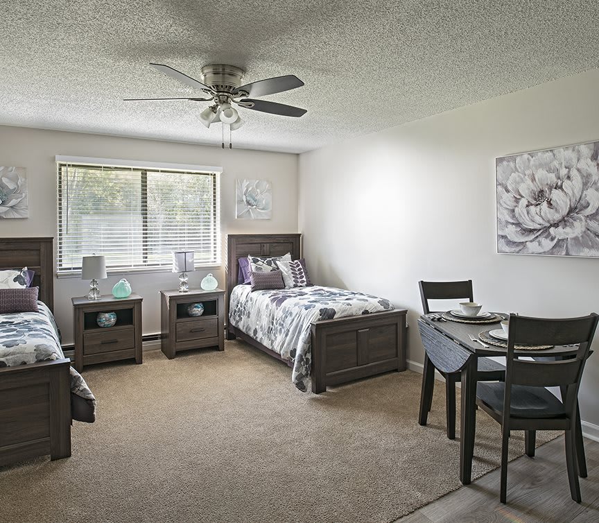 A spacious bedroom with two twin beds, each accompanied by a nightstand. The room features a large window for natural light, a ceiling fan, and a small dining table with chairs.