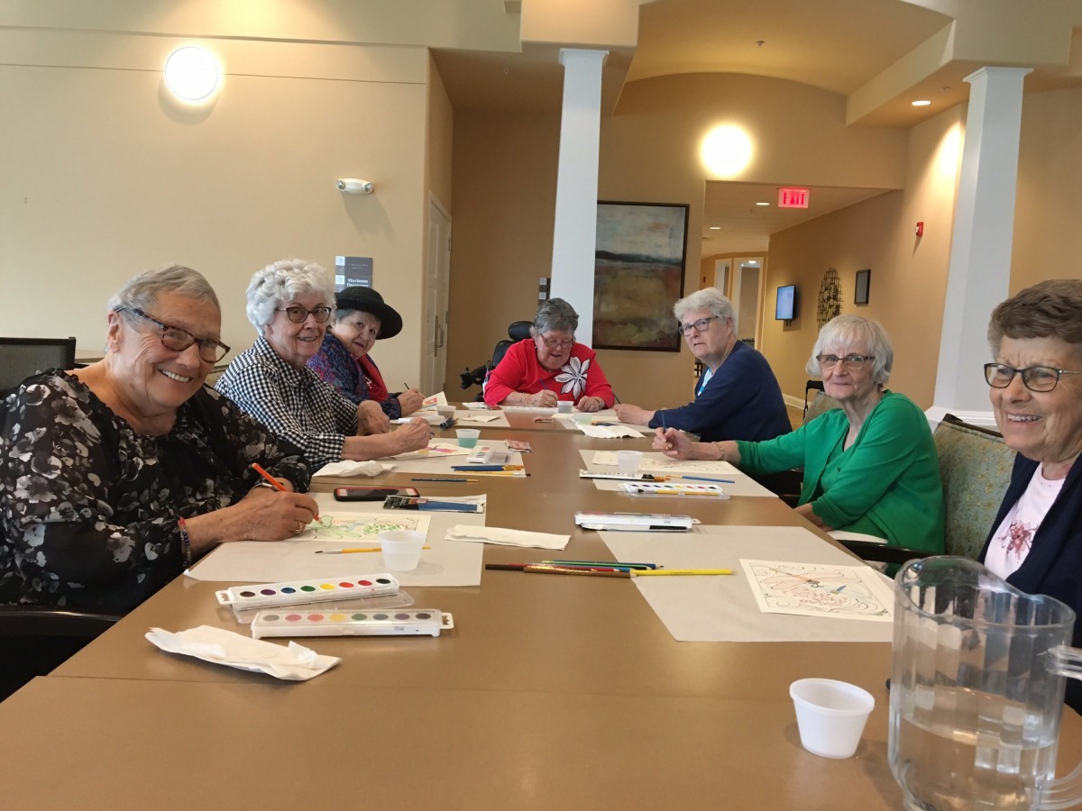 A group of seniors participating in an art class, seated around a table with painting supplies.